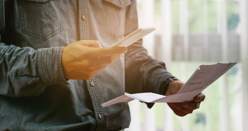 Mit niedrigeren Fixkosten ins neue Jahr – Tipps zum Geld Sparen 1 Die Hände eines Mannes, der in einer Hand ein Smartphone und in der anderen einen Zettel hält, Stichwort Fixkosten.
(c) AdobeStock