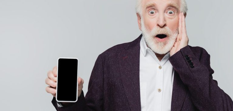 Ein alter Mann mit Vollbart hält mit offenem Mund erstaunt ein Smartphone in die Kamera. (c) AdobeStock