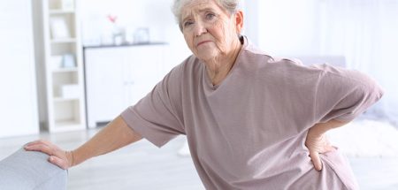 Eine alte Frau, die sich mit einer Hand auf einer Couch abstützt und mit der anderen auf ihren schmerzenden Rücken greift. (c) AdobeStock