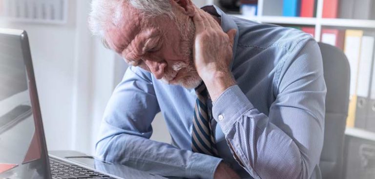 Ein älterer Mann mit Bart greift sich an einem Schreibtisch sitzend vor Schmerzen mit der Hand in den Nacken. (c) AdobeStock