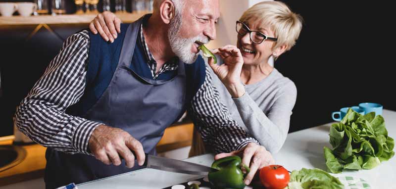 Eine alte Frau, die einem alten Mann, der einen Paprika schneidet, ein Stück davon in den Mund gibt. (c) AdobeStock