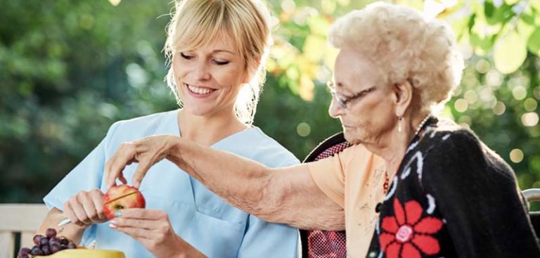 Eine alte Frau nimmt ein Stück Apfel, das eine junge Pflegerin neben ihr aufschneidet. (c) AdobeStock
