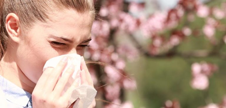 Eine Frau, die sich die Nase putzt aufgrund einer Allergie; im Hintergrund ein blühender Baum. (c) AdobeStock