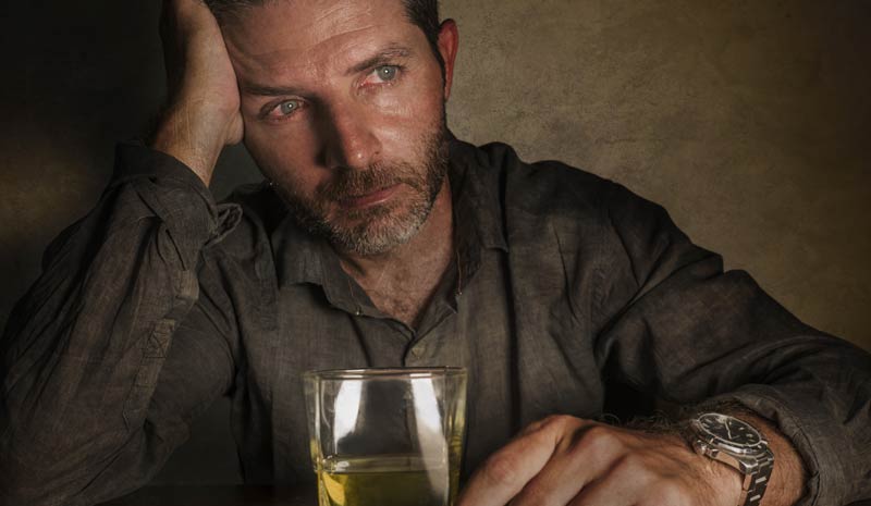 Ein trauriger Mann, der seinen Kopf an seine Hand stützt und vor sich ein Glas Whiskey stehen hat.
(c) AdobeStock