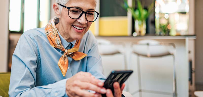 Eine ältere Frau mit Brille schaut lächelnd auf ihr Smartphone. (c) AdobeStock