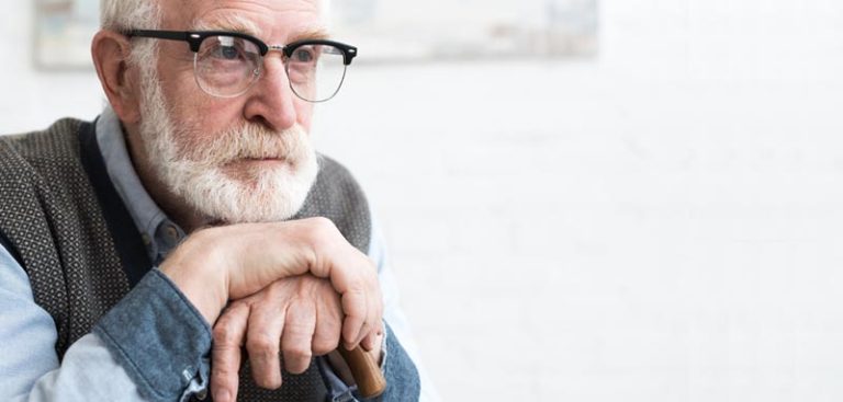 Ein alter Mann mit Vollbart und Brille mit dem Kinn auf seine einen Gehstock haltenden Hände gestützt. (c) AdobeStock