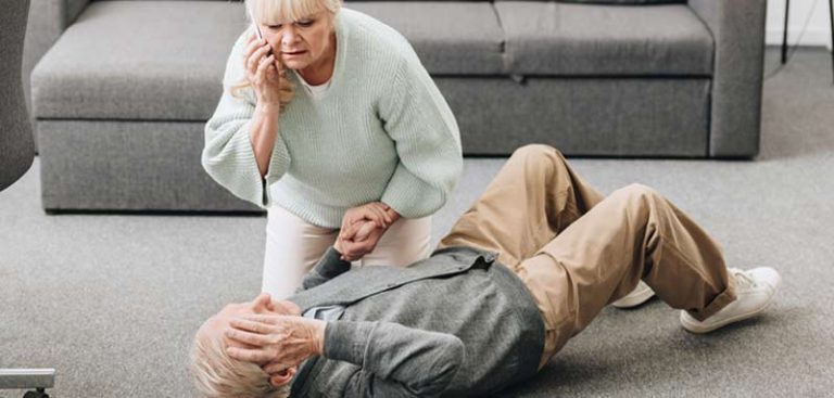 Eine ältere Frau, die neben einem auf dem Boden liegenden älteren Mann kniet, seine Hand hält und nach Hilfe telefoniert. (c) AdobeStock