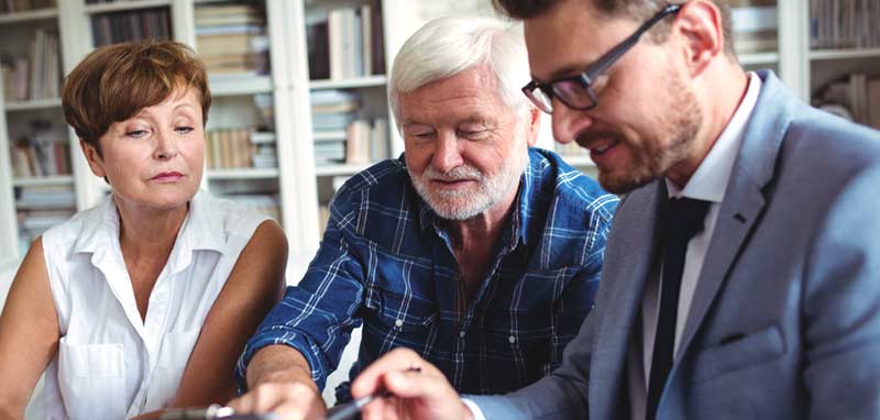 Ein Finanzberater mit einem älteren Ehepaar in deren Wohnzimmer. (c) AdobeStock