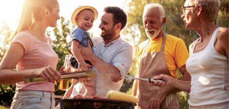 Eine Frau, ein Mann mit einem kleinen Kind am Arm und ein älteres Paar um einen Griller. (c) AdobeStock