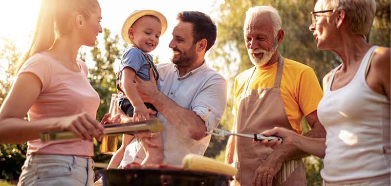 Eine Frau, ein Mann mit einem kleinen Kind am Arm und ein älteres Paar um einen Griller. (c) AdobeStock