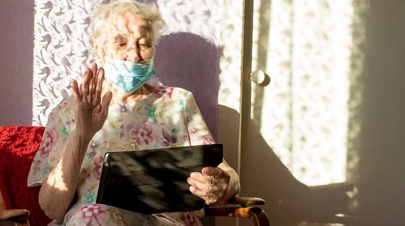 Eine alte Frau mit Mund-Nasen-Schutz, die ein Tablet hält und zur Begrüßung winkt.
(c) AdobeStock