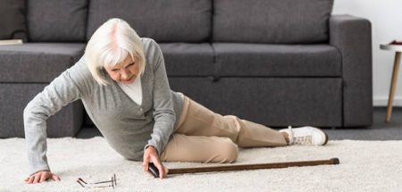 Eine alte Frau liegt nach einem Sturz auf einem Teppich vor einer Couch. (c) AdobeStock