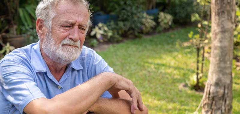 Ein älterer Mann mit Vollbart sitzt auf einer Bank und schaut ins Leere. (c) AdobeStock