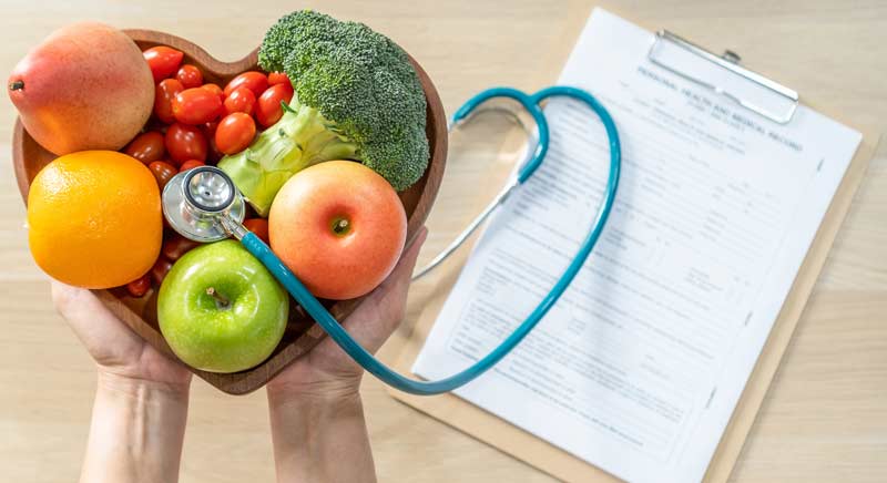 Jemand hält eine herzförmige Schüssel mit Obst und Gemüse und einem Stethoskop, darunter liegt ein Fragebogen, Stichwort Cholesterin.
(c) AdobeStock