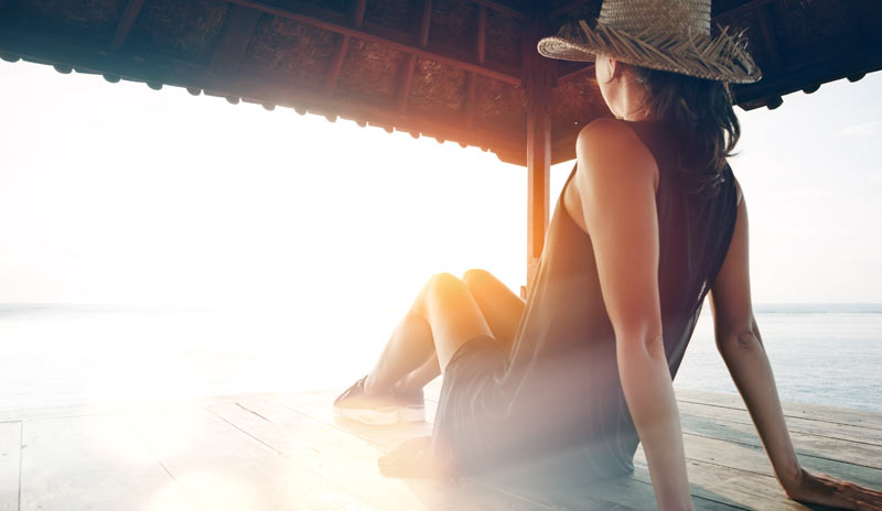 Eine Frau mit Sonnenhut auf einem Sonnendeck an einem See unter einem Sonnenschutz.
(c) AdobeStock