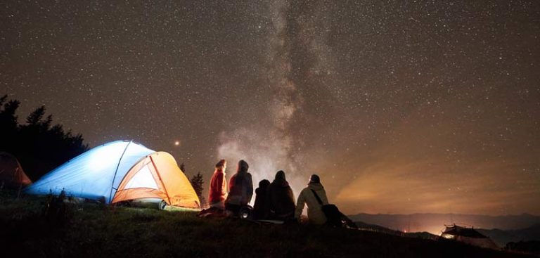 Ein beleuchtetes Zelt in der freien Natur in der Nacht unter Sternenhimmel, davor vier Personen um ein Lagerfeuer. (c) AdobeStock