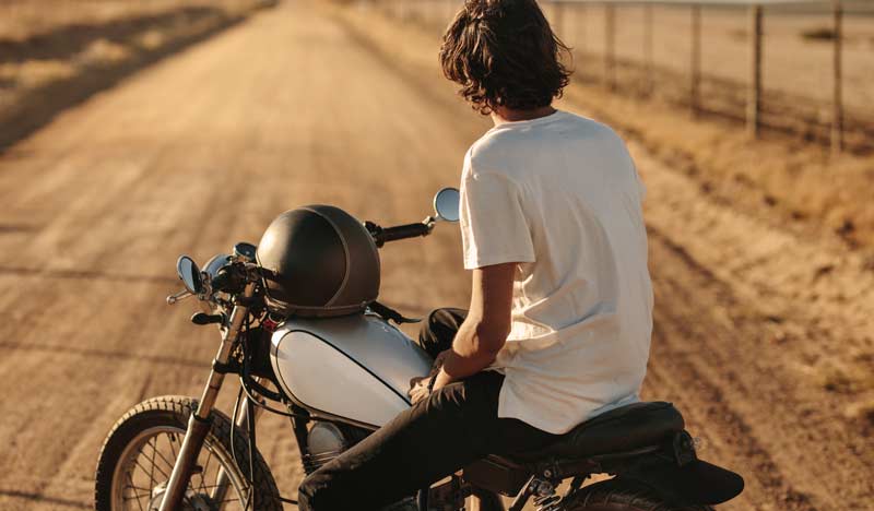 Ein Jugendlicher sitzt auf einem Motorrad der 1960er auf einer unasphaltierten Landstraße und schaut Richtung Horizont.
(c) AdobeStock