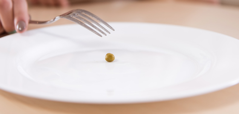 Die Hand einer Frau, die mit einer Gabel eine einzelne Erbse auf einem Teller essen möchte. (c) AdobeStock