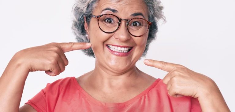 Ein lachende Frau, die mit ihren Zeigefingern auf ihre weißen Zähne zeigt. (c) AdobeStock