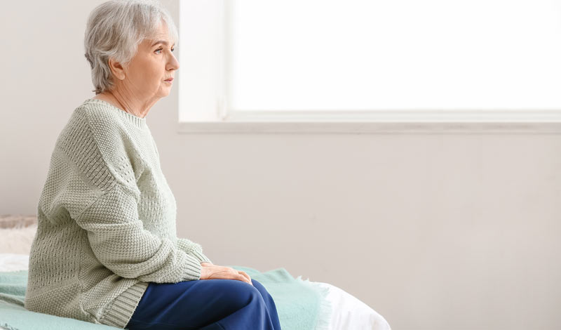 Eine ältere Frau sitzt auf ihrem Bett und schaut ins Leere, Stichwort Alzheimer.
(c) AdobeStock