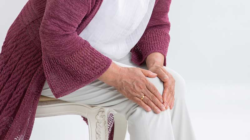 Die Hände einer alten Frau, die sich auf einem Sessel sitzend auf ihr schmerzendes Knie greift, Stichwort Gelenksbehandlung.
(c) AdobeStock