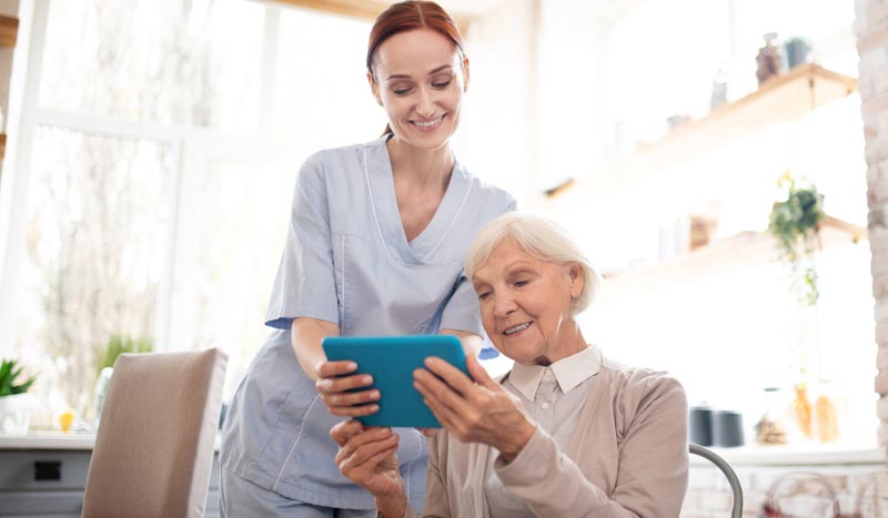 Eine Pflegerin zeigt einer alten Frau etwas auf einem Tablett, Stichwort Community Nursing.
(c) AdobeStock