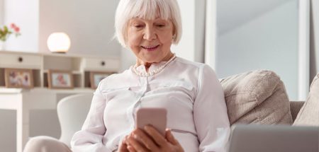 Eine alte Frau sitzt auf einer Couch mit einem Smartphone in der Hand. (c) AdobeStock