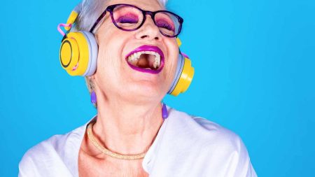Eine ältere Frau mit Brille, purpuren Lippen und Liedschatten und gelben Kopfhörern singt laut mit zur Musik, die sie hört. (c) AdobeStock