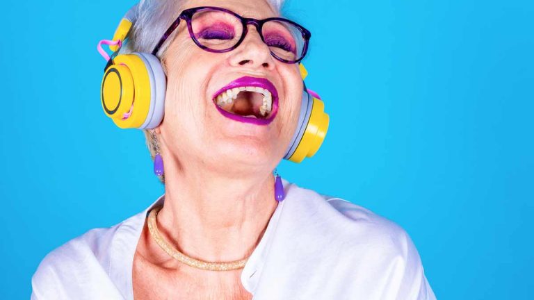 Eine ältere Frau mit Brille, purpuren Lippen und Liedschatten und gelben Kopfhörern singt laut mit zur Musik, die sie hört. (c) AdobeStock
