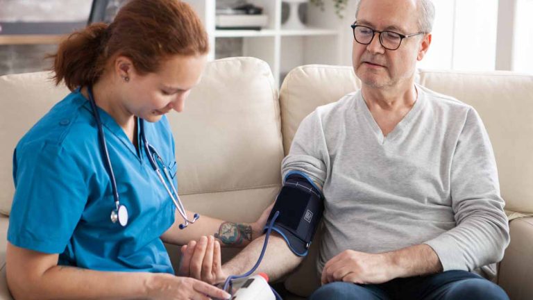Eine Pflegerin, die einem Mann zu Hause den Blutdruck misst. (c) AdobeStock