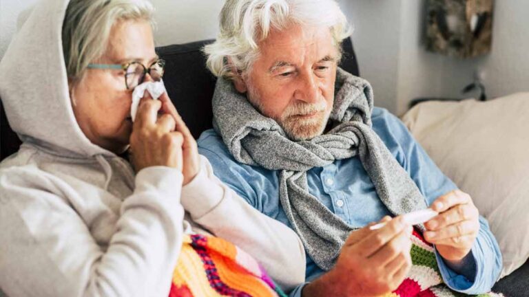 Ein älteres Paar krank mit Grippe auf einem Sofa; die Frau putzt sich die Nase, der Mann schaut auf das Fieberthermometer. (c) AdobeStock