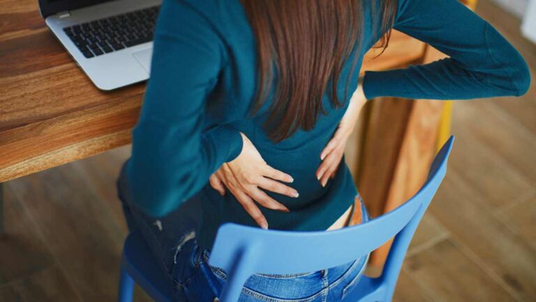 Eine Frau, die auf einem Sessel sitzt und sich vor Schmerzen mit beiden Händen an den unteren Rücken greift. (c) AdobeStock