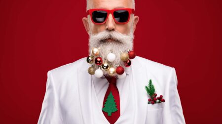 Ein Mann mit weißem Vollbart, roter Sonnenbrille, Weihnachtskugeln im Bart und einer roten Krawatte mit grünem Tannenbaum. (c) AdobeStock