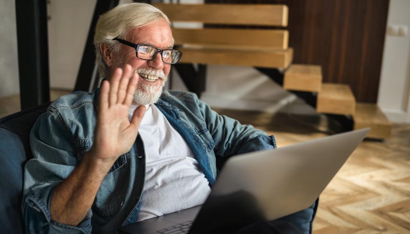 Ein älterer Mann mit Bart und Brille sitzt in einem Sessel und winkt in seinen Laptop, Stichwort Fernbeziehung.
(c) AdobeStock