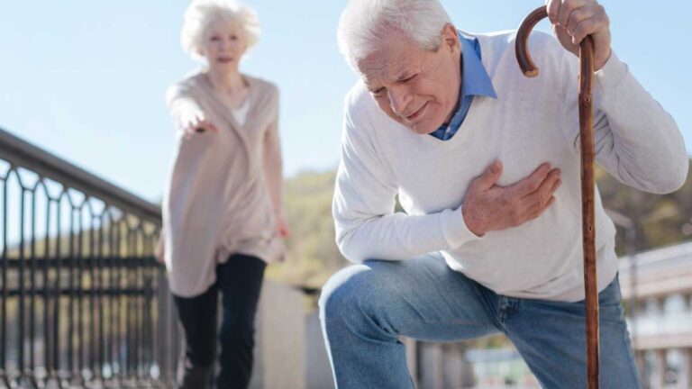 Ein alter Mann kniet sich, auf einem Gehstock gestützt, vor Schmerzen in der Herzgegend nieder, dahinter eilt eine alte Frau zu Hilfe. (c) AdobeStock