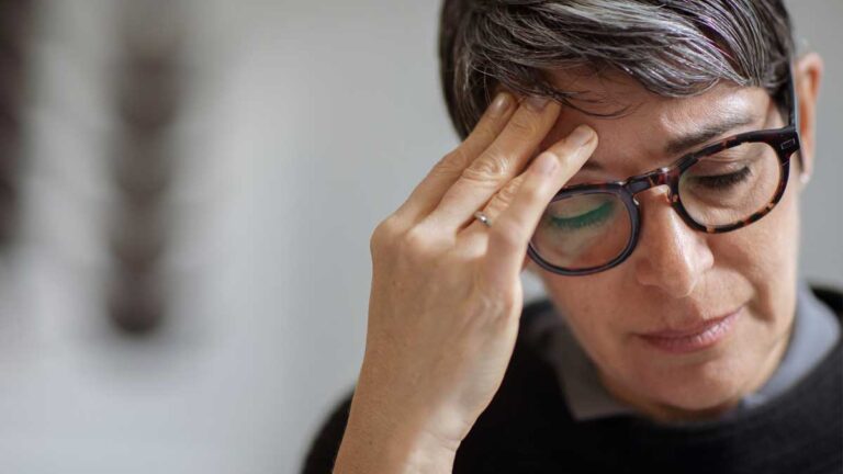 Eine Frau mit Brille, die sich mit der Hand an den Kopf greift wegen Kopfschmerzen. (c) AdobeStock