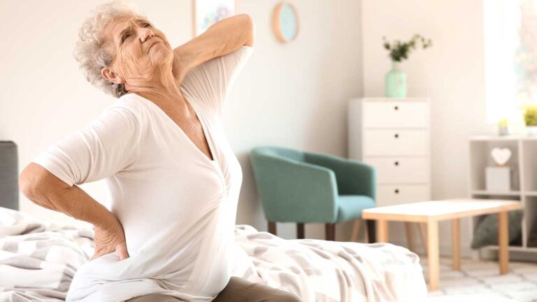 Eine ältere Frau, die sich auf einem Bett sitzend vor Schmerzen den Rücken und Nacken hält. (c) AdobeStock