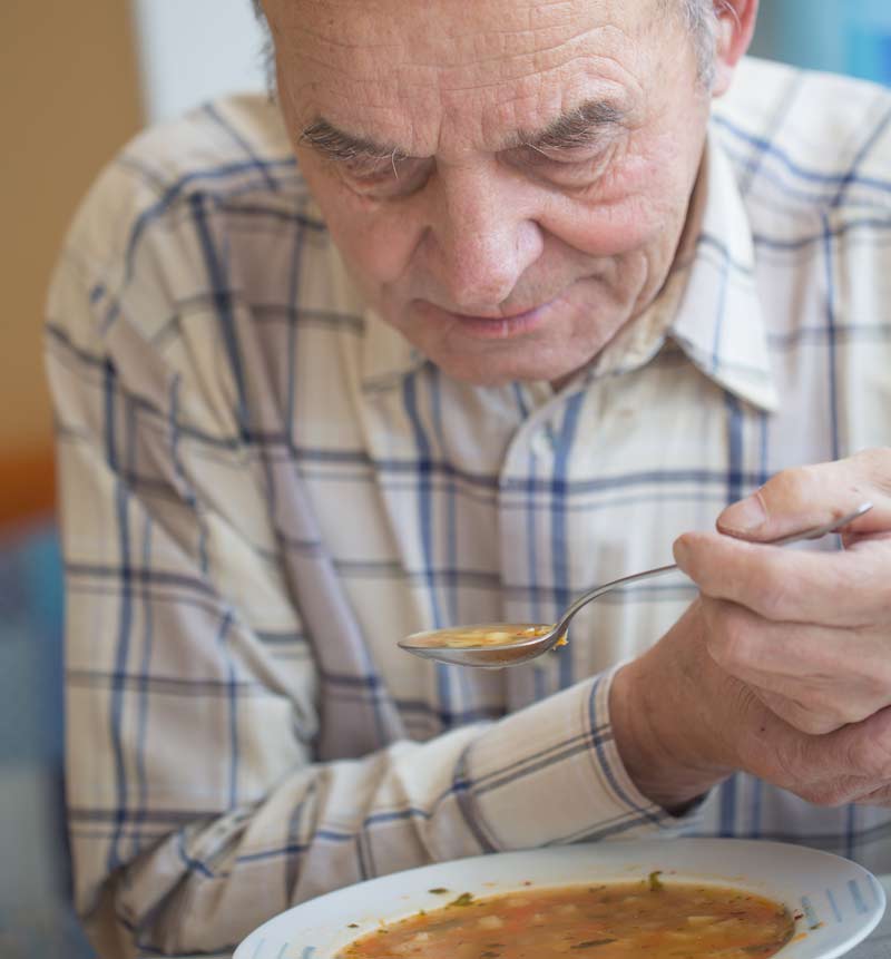 Ein Mann, der sich beim Essen einer Suppe mit einer Hand die andere zitternde hält, Stichwort Morbus Parkinson.
(c) AdobeStock