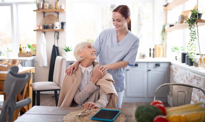 Eine Pflegerin, die einer alten Frau, die an einem Tisch sitzt, die Hände auf die Schultern legt, Stichwort Pflege neu denken.
(c) AdobeStock