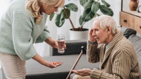 Eine Frau gibt einem sitzenden alten Mann Medikamente. (c) AdobeStock