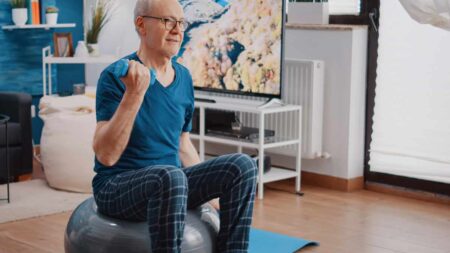 Ein älterer Mann zu Hause auf einem Gymnastikball sitzend beim Training mit Kurzhanteln. (c) AdobeStock