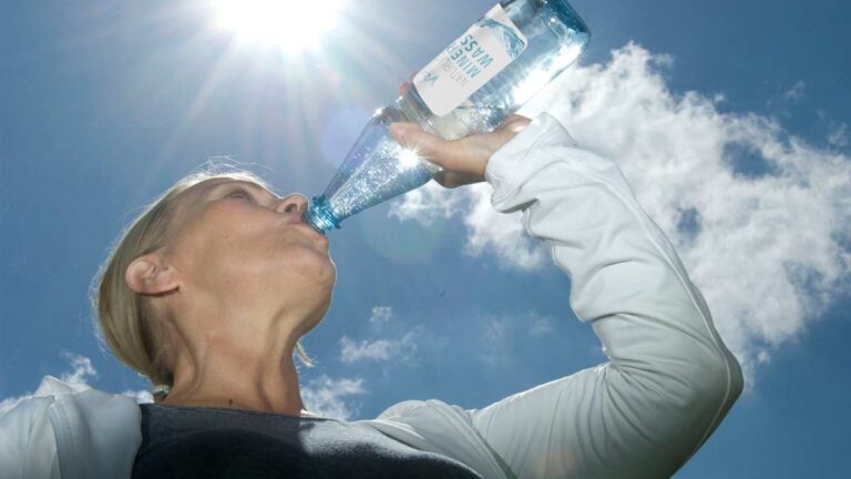 Eine Frau, die aus einer Mineralwasserflasche trinkt, über ihr die Sonne. (c) Informationszentrale Deutsches Mineralwasser