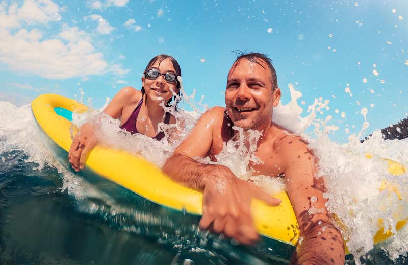 Wie wirkt sich »das Reisen« auf unsere psychische Gesundheit aus? 1 Ein Mann und ein Mädchen im Meer auf einem Surfbrett.
(c) AdobeStock