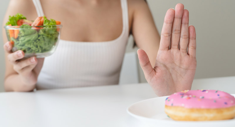 Die Hände einer Frau, die in einer eine Schüssel mit Salat hält und mit der anderen "Stop" in Richtung eines Dougnuts zeigt.
(c) AdobeStock
