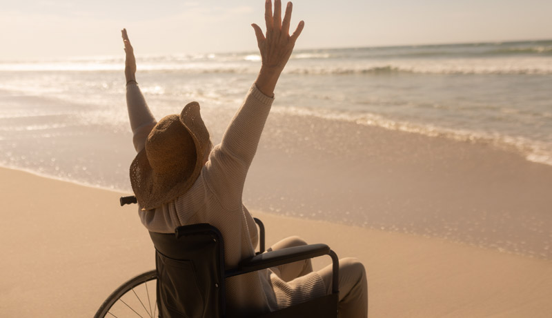 Menschen mit Behinderung erleben oft einen Urlaub mit Hindernissen 2 Eine Frau im Rollstuhl am Strand mit in die Höhe gestreckten Armen.
(c) AdobeStock