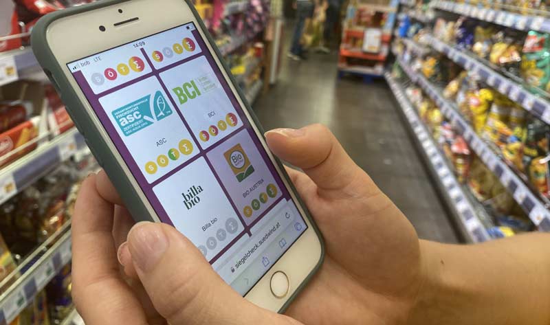 Screen eines Handys, das ein Mann in einem Supermarkt hält, mit dem Gütesiegel-Check Online Tool.
(c) Südwind
