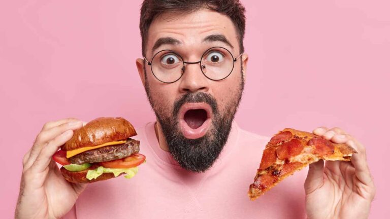 Ein Mann mit aufgerissenem Mund und Augen hält einen Burger und ein ein Stück Pizza. (c) AdobeStock