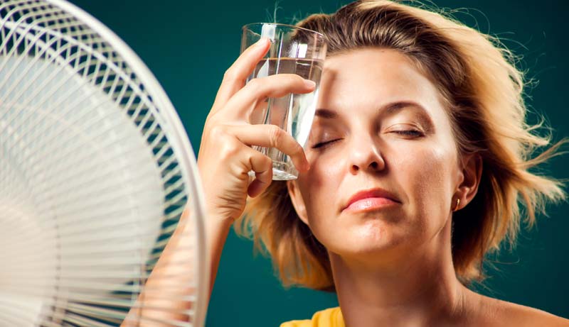 Eine Frau sitzt vor einem Ventilator und hält sich ein Wasserglas zur Kühlung an die Stirn.
(c) AdobeStock