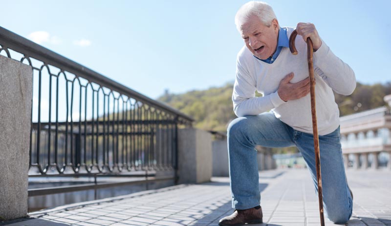 Ein alter, auf einem Bein kniender Mann, der sich mit einer Hand vor Schmerzen an sein Herz greift und mit der andern auf einem Gehstock abstützt.
(c) AdobeStock