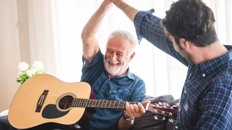 Ein älterer Mann mit Gitarre gibt einem Gitarrenlehrer high five. (c) AdobeStock